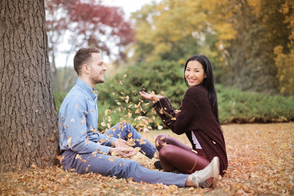 Side view of cheerful couple in casual clothes sitting in park and having fun while scattering dry leaves on autumn day